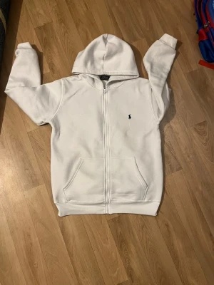 Vit zip hoodie från Polo Ralph Lauren - Klassisk vit hoodie med dragkedja från Polo Ralph Lauren. Har huva, två fickor framtill och den ikoniska lilla svarta loggan broderad på bröstet. Tillverkad i mjukt material som känns skönt mot huden. Perfekt för en avslappnad och stilren look.