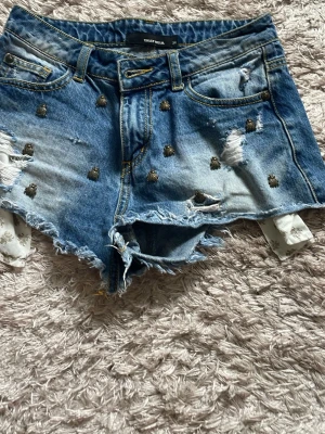 Blå jeansshorts med nitar från Truly Well - Säljer ett par blå jeansshorts från Truly Well med slitna detaljer, rå kant och små nitar framtill. Shortsen har klassisk femficksdesign och gul kontrastsöm. Perfekta för sommarens alla äventyr!