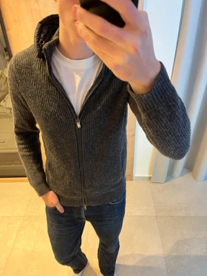 Mörkgrå stickad cardigan med huva från Massimo Dutti - Riktigt snygg mörkgrå stickad cardigan jacka i väldigt fint skick. Notera att den inte är stickad på ryggen (se bild 2).                                              ———————————————————————————                                               📐| Storlek: M                                                           👖| Märke: Massimo Dutti                                        🔎 | Skick: Mycket Bra                                              ✍🏻 | Skriv om du har några frågor
