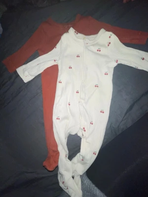 Mini market pyjamas  - Säljer två mysiga pyjamasar för barn, en i vit med små röda körsbär och en i roströd. Båda har långa ärmar och ben med fötter, samt dragkedja framtill för enkel påklädning. Perfekta för sköna hemmakvällar eller sovstunder.