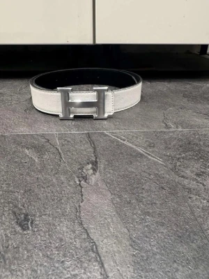  vitt skärp med H-spänne - Stilrent vitt skärp från med ikoniskt H-format spänne i silverfärgad metall. Bältet är gjort i skinn och har en klassisk, tidlös design som passar till många olika outfits.