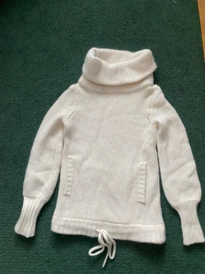 Vit polotröja från Reiss ull 70% och alpaca 20% - Säljer en krämvit stickad polotröja från Reiss med bred polokrage och dragsko i nederkanten. Tröjan har långa ärmar, ribbstickade muddar och två dekorativa fickor framtill. Perfekt för kalla dagar och riktigt mysig att bära.