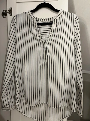 Vit randig blus  - Vit blus med svarta vertikala ränder från.Blusen har v-ringning med knappdetalj och långa ärmar med manschett. Tillverkad i ett lätt och mjukt tyg som ger en avslappnad och stilren look. 