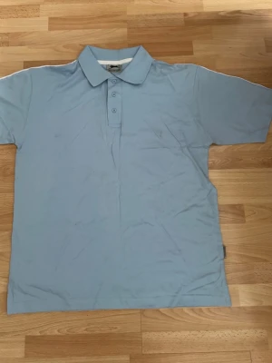 Ny. Ljusblå pikétröja från Slazenger - Ljusblå pikétröja från Slazenger med klassisk krage och tre knappar framtill. Tröjan har korta ärmar och en vit rand längs axlarna på baksidan. Perfekt för en sportig och avslappnad stil. Bomull 100%