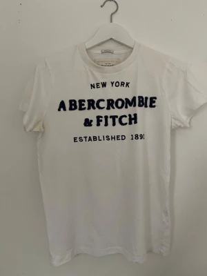 Vit t-shirt Abercrombie & Fitch - Vit t-shirt från Abercrombie & Fitch med blå text på bröstet och klassisk rund hals. Modellen är 'Muscle' och har korta ärmar. Perfekt för dig som gillar en enkel och stilren look med tydlig logga. Gjord i mjuk bomull för skön komfort. Har lite gula fläckar men eftersom att det är en muscle fit så syns det inte när den är på. Jätte mjukt material.