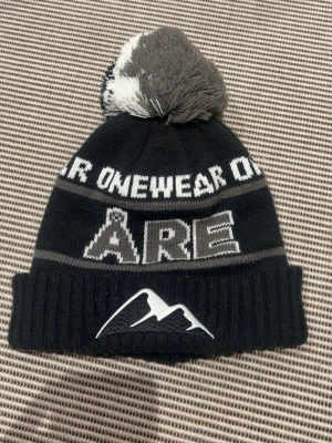 Svart Åre mössa med tofs - Säljer en svart stickad mössa med grå och vit text 'ÅRE' och bergmotiv framtill. Mössan har en stor tofs i grått och vitt på toppen och vikt kant. Perfekt för kalla dagar och snygg till vinterlooken. Använd 1 gång.