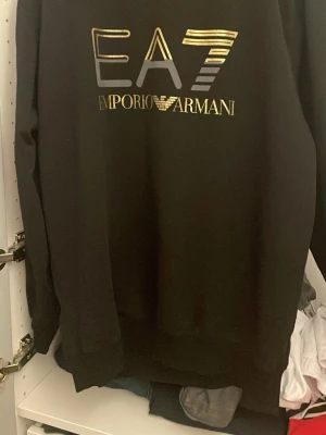 Svart EA7 Emporio Armani hoodie - Snygg svart hoodie från EA7 Emporio Armani med stor guldfärgad logga och text på bröstet. Storlek XXL står det i tröjan  men skulle säga att den passar L 