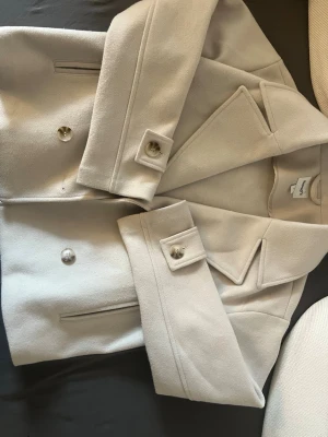 Kort beige kappa från Kappahl  - Stilren kort kappa i beige från Kappahl med breda slag, dubbelknäppning och dekorativa knappar på ärmsluten. Kappan har raka fickor framtill och är gjord i ett mjukt, ull-liknande material. Perfekt för dig som gillar minimalistisk och trendig stil.