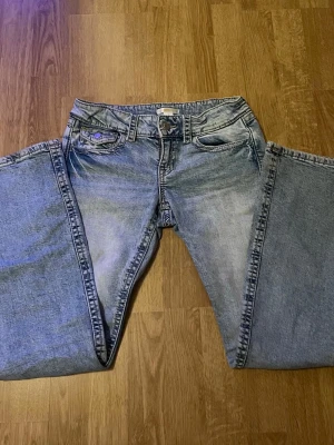 Ljusblå bootcut jeans med fickdetaljer - Säljer ett par ljusblå bootcut jeans med snygga fickor med lock och knapp bak. Klassisk femficksmodell i mjuk denim, perfekta till en avslappnad stil. Passar dig som gillar vintagevibbar och bredare ben. Fråga vad ni vill och pris kan diskuteras!