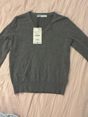 Grå stickad tröja från Zara - Snygg grå stickad tröja från Zara med V-ringning. Tröjan har ribbade muddar vid ärmslut och nederkant. Tröjan är liten i storlek men mycket fin. Helt oanvänd och lappen är kvar. 