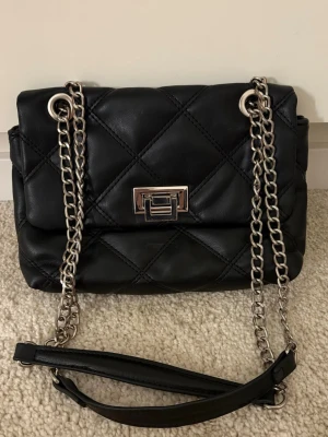 Svart handväska med kedja - Snygg svart axelväska i quiltat konstläder med silverfärgad kedjerem och metallspänne framtill. Väskan har en stilren, rektangulär form och detaljer som ger en trendig look. 