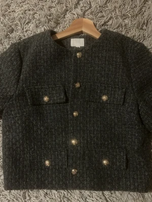 Mörkgrå bouclé cardigan med guldknappar - Stilren mörkgrå cardiganjacka i bouclé med rund halsringning och dekorativa guldknappar framtill. Fyra fickor och långa ärmar. Perfekt för dig som gillar klassisk och trendig stil.