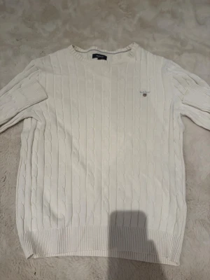 Vit kabelstickad tröja från GANT - Kabelstickad vit tröja från GANT med rund halsringning och diskret logga på bröstet. Tröjan har ribbade muddar vid ärmslut och nederkant, klassisk passform och är tillverkad i mjukt material som känns skönt mot huden.