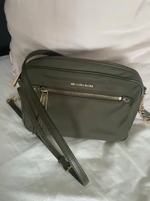 Olivgrön axelväska Michael Kors - Snygg olivgrön axelväska från Michael Kors i slätt skinn med guldfärgade detaljer och kedja på axelremmen. Väskan har en dragkedja framtill, justerbar rem och stilren form. Perfekt för dig som vill ha en trendig och praktisk väska med exklusiv känsla. Fint skick förutom dom små smink fläckarna.