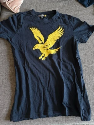 Blå t-shirt med gul örntryck - Cool mörkblå t-shirt från Lyle & Scott med stort gult örntryck på bröstet. Klassisk passform och rund halsringning, perfekt för dig som gillar sportiga och stilrena plagg. Materialet är mjuk bomull som känns skönt mot huden.