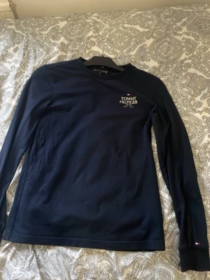 Mörkblå långärmad Tommy Hilfiger - Snygg mörkblå långärmad tröja från Tommy Hilfiger med diskret logga på bröstet och liten flagga på ärmslutet. Tröjan har rund halsringning och är tillverkad i mjukt bomullsmaterial. Perfekt för en clean och stilren look.