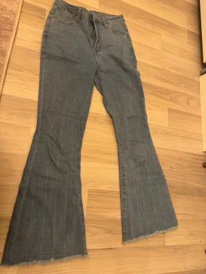 Blå utsvängda jeans med rå kant - Säljer ett par blå jeans med utsvängda ben och råa, fransiga benslut. Jeansen har klassisk femficksmodell och hög midja. Materialet är denim i bomull och de har subtila vertikala sömmar längs benen för en cool vintagekänsla.