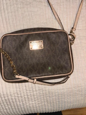 Brun axelväska från Michael Kors - Snygg brun axelväska från Michael Kors med klassiskt MK-mönster, guldfärgad kedjedetalj på axelremmen och beigea kanter. Väskan har en rektangulär form och en metallbricka med logga på framsidan. Perfekt för dig som gillar stilrena accessoarer.