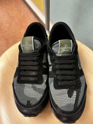 Valentino Garavani camo sneakers - Snygga sneakers från Valentino Garavani med svart och grått camomönster. Ovandelen är i mesh och mocka, med svarta skosnören och robust sula. Perfekta för dig som vill ha en unik och trendig look.