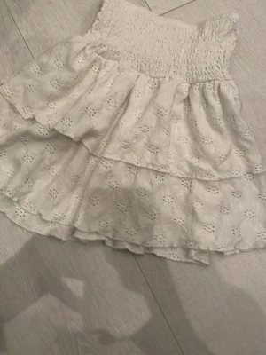 Vit volangkjol med broderade detaljer - Supersöt vit kortkjol med volanger och broderade blommor. Kjolen har en smockad midja som ger en bekväm passform och är perfekt för varma dagar. Materialet känns lätt och luftigt, vilket gör den extra skön att bära.