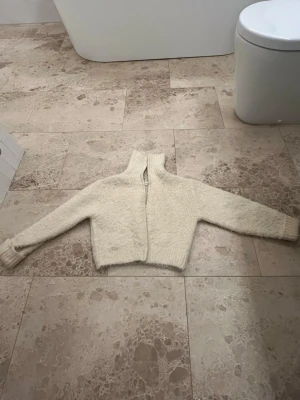 Krämvit fluffig zip-kofta - Mysig krämvit kofta med fluffig textur och dragkedja framtill. Hög krage och långa ärmar med ribbade muddar. Perfekt för lager-på-lager och ger en cool, avslappnad look. Materialet känns mjukt och varmt.