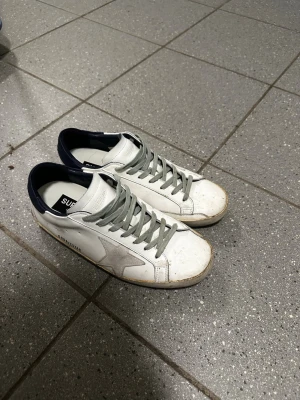Golden goose - Snygga vita sneakers från golden goose med grå snörning och mörkblå häl. Skorna har en klassisk låg siluett och en stjärndetalj på sidan. Tillverkade i läder med gummisula, perfekta för en clean och stilren look. Priset är ej hugget i sten så varm inte rädda att ställa frågor 😁