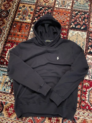 Mörkblå hoodie från Polo Ralph Lauren - Klassisk mörkblå hoodie från Polo Ralph Lauren med den ikoniska broderade loggan på bröstet. Hoodien har huva, ribbade muddar och magficka. Tillverkad i mjuk bomullsblandning som känns skön mot huden. Perfekt för en avslappnad och stilren look.