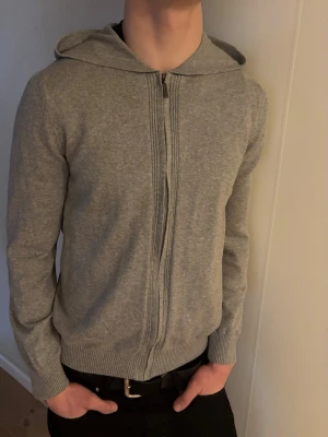 Grå zip hoodie - Säljer en stilren ljusgrå hoodie med dragkedja framtill och ribbade muddar. Hoodien är storlek mnoch sitter såhär på 188cm. Mycket bra skick!