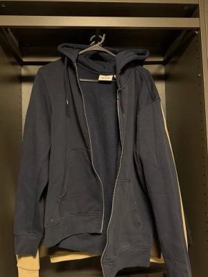 Mörkblå hoodie från Weekday - Mörkblå hoodie från Weekday med dragkedja framtill, två fickor och justerbar huva med snören. Tillverkad i mjuk bomullsblandning och har ribbade muddar vid ärmslut och nederkant. Perfekt för en avslappnad och stilren look. OBS!! DRAGKEDJAN FUNKAR INTE!