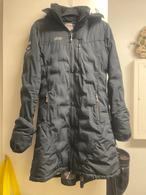 Marinblå knälång dunjacka från Uhip - Säljer en marinblå  dunjacka från Uhip med hög krage och flera praktiska dragkedjor. Jackan har ett sportigt snitt, diskreta reflexdetaljer på ärmen och är tillverkad för att hålla dig varm under kalla vinterdagar. Perfekt för dig som gillar funktionella kläder. Fint skick, däremot lagad vid ena armen (armhålan) se bild 5. Inget som påverkar kvaliteten, en mycket bra jacka! 