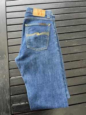Blå jeans från Nudie  - Säljer ett par klassiska blå jeans från Nudie med raka ben och snygga kontrastsömmar. Jeansen har fem fickor, gul brodyr på bakfickan och patch i läder bak i midjan. Jeansen är i fint skick och storlek W29 L34 pris går att diskutera