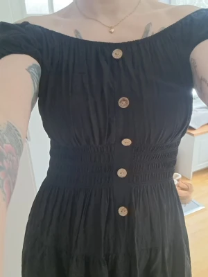 Svart offshoulder klänning med knappar - Svart klänning med offshoulder-modell och träfärgade knappar framtill. Klänningen har smockad midja som ger en snygg siluett och kort ärm. Perfekt för dig som gillar en feminin och trendig stil. Mjuk luftig och perfekta på sommaren