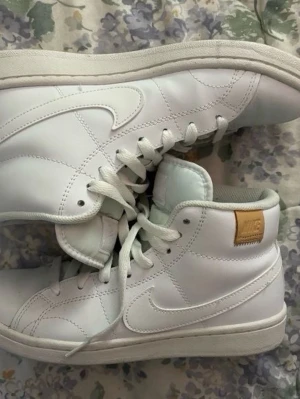 Vita Nike high top sneakers i läder - Snygga vita high top sneakers från Nike i läder med klassisk swoosh på sidorna och beige Nike-märke på hälen. Skorna har vita snören och en vit sula, samt diskreta sömdetaljer och plösen är någorlunda turkos. De är använda en gång 
