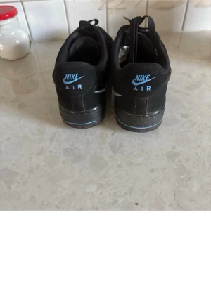 Svarta Nike Air Force 1 sneakers - Säljer ett par klassiska svarta Nike Air Force 1 sneakers med blå detaljer på hälen och sulan. Skorna har lågt skaft, snörning och är tillverkade i läder med den ikoniska swoosh-loggan på sidan. Perfekta för dig som gillar streetwear och tidlös stil.
