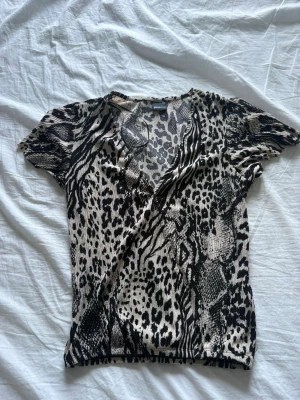 Leopardmönstrad topp från Gina Tricot - Snygg kortärmad topp från Gina Tricot i beige och svart med leopardmönster. Toppen har v-ringning och är tillverkad i ett stretchigt syntetmaterial som sitter tight. Perfekt för dig som vill sticka ut med ett djurmönster.