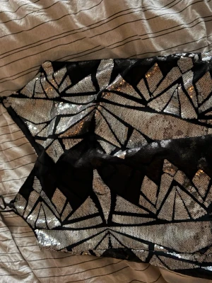 Svart och silver glittrig linnetopp - Säljer en svart linnetopp med tunna axelband och geometriskt mönster i silverpaljetter. Toppen har en glittrig look och är perfekt till fest!