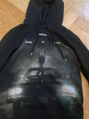 Svart hoodie Dondup med biltryck - Svart hoodie från Dondup med stor grafisk print framtill som visar en siluett av en person på en bil. Texten 'MADE IN ITALY' syns ovanför trycket. Hoodien har dragsko i huvan och ribbade muddar. Perfekt för dig som gillar streetwear och unika prints.
