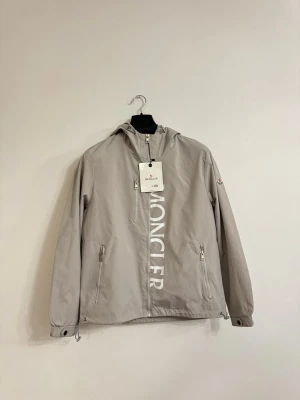  Moncler windbreaker i grå  - Moncler Windbreaker i ljusgrå. Helt ny med tagg kvar, aldrig använd. Riktigt clean färg och perfekt nu till våren/sommaren.  Storlek M.  