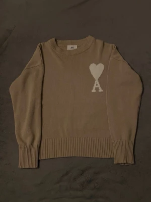 Beige stickad tröja från Ami Paris - Snygg beige stickad tröja från Ami Paris med vitt hjärta och A-logga på bröstet. Tröjan har rund halsringning, långa ärmar och breda ribbade muddar vid ärmslut och nederkant. Perfekt för en stilren och trendig look.