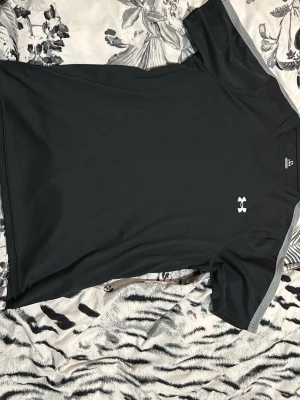 Svart tränings-tshirt från Under Armour - Säljer en svart tränings-tshirt från Under Armour med grå detaljer på axlarna och vitt logotryck på bröstet. Tillverkad i ett snabbtorkande funktionsmaterial som passar perfekt till gymmet eller löprundan.