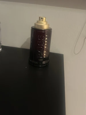 BOSS The Scent Elixir parfym - En elegant parfym från Hugo Boss, The Scent Elixir. Flaskan är mörkröd med guldfärgat lock och detaljer. Den har en stilren, cylindrisk form och utstrålar lyxig känsla. Perfekt för dig som gillar exklusiva dofter och snygg design. Ingen parfym inuti och locket är borta