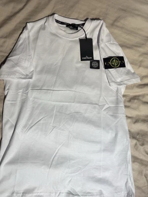 Vit Stone Island t-shirt - Snygg vit t-shirt från Stone Island med klassisk logopatch på ärmen. T-shirten har rund hals och korta ärmar, tillverkad i mjuk bomull för en clean och stilren look. Perfekt för dig som gillar streetwear och vill ha något enkelt men ändå exklusivt.
