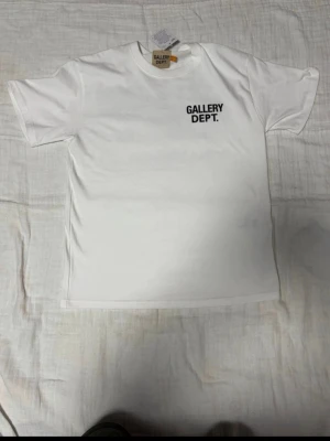 Vit t-shirt från Gallery Dept. - Snygg vit t-shirt från Gallery Dept. med svart logotyptryck på bröstet. Klassisk rund hals och korta ärmar. Tillverkad i mjuk bomull som känns skön mot huden. Perfekt för dig som gillar enkel och stilren streetwear.