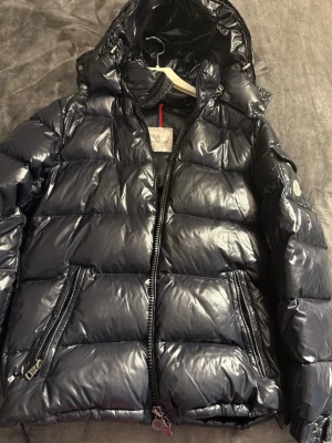Moncler Maya - Snygg svart dunjacka från Moncler med glansig finish och huva. Jackan har två dragkedjeförsedda fickor framtill och Moncler-logga på ärmen. Perfekt för kalla vinterdagar och har en klassisk puffer-modell. Storlek S kan passa XS och M