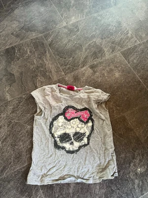 Grå t-shirt med döskalletryck - Säljer en grå t-shirt med ett stort döskalletryck framtill och en röd detalj med texten 'Monster High' ovanför motivet. T-shirten har rund hals och korta ärmar, perfekt för dig som gillar edgy och cool stil. (Plus frakt) 