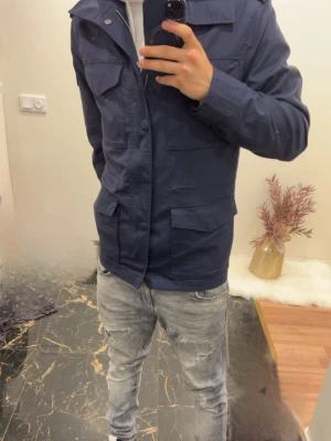 Pierre cardin Mörkblå utilityjacka med fickor - Snygg mörkblå utilityjacka med fyra stora fickor framtill och dragkedja samt knappar. Jackan har en rak passform och klassisk krage, perfekt för en stilren och avslappnad look. Materialet ser ut att vara bomull eller bomullsblandning. Nästan ny, inga skador! Storlek M . Ordinarie pris 3200kr+ 