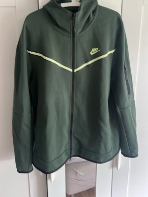 Grön Nike hoodie med dragkedja XL - Snygg grön hoodie från Nike med hel dragkedja och huva. Har en ljusgrön detalj över bröstet och Nike-logga på vänster sida. Ficka med dragkedja på ärmen. Perfekt för en sportig och avslappnad stil.