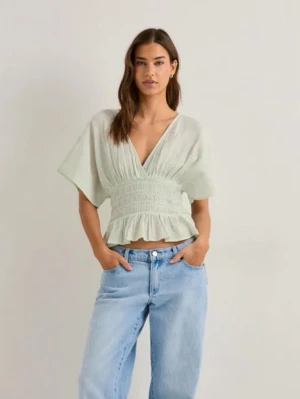 Ljusgrön blus med v-ringning - Superfin ljusgrön blus med djup v-ringning, korta vida ärmar och smockad midja. Toppen har volangkant nedtill och är tillverkad i ett lätt och luftigt bomullstyg. Perfekt att styla med jeans eller kjol för en trendig look.