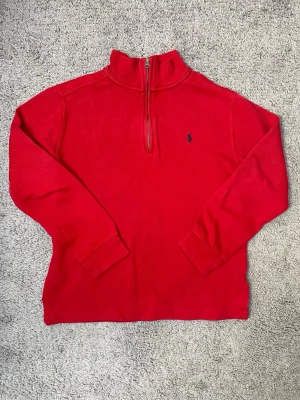 Röd halvzip från Polo Ralph Lauren - Riktigt snygg röd tröja från Polo Ralph Lauren med halv dragkedja och broderad logga på bröstet. Tröjan har hög krage och långa ärmar, perfekt för lager-på-lager. Klassisk och stilren design som funkar året runt.