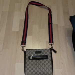 Axelväska från GG med monogram - Snygg axelväska från GG med klassiskt monogrammönster. Väskan har en justerbar axelrem i rött och blått samt en praktisk dragkedja framtill. Insidan är fodrad med GG-logotypen. Pris kan diskuteras vid snabb affär.